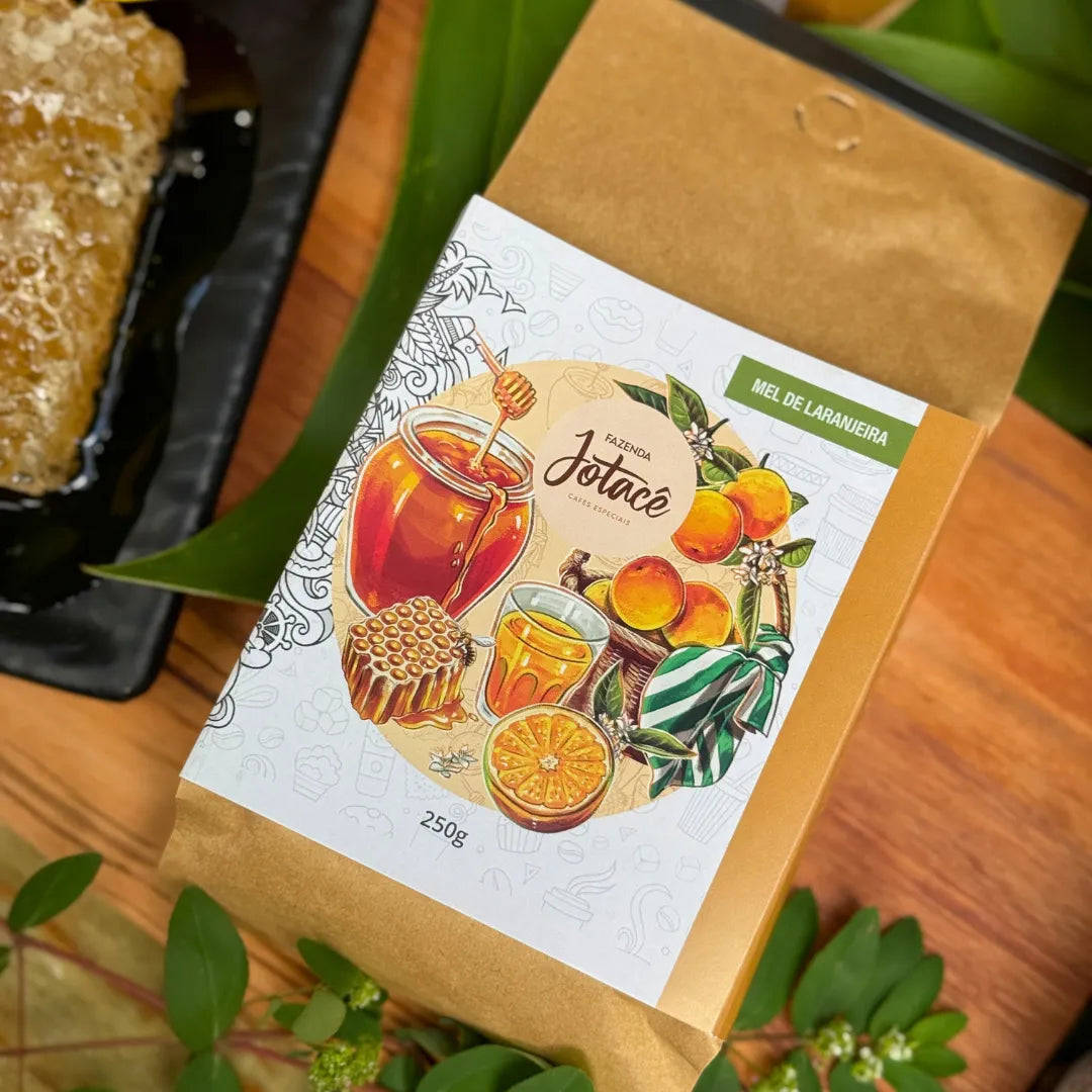 Café especial Mel de Laranjeira da Fazenda Jotacê sobre mesa de madeira, com laranja cortada e favo de mel ao lado, reforçando as notas doces e cítricas do café.
