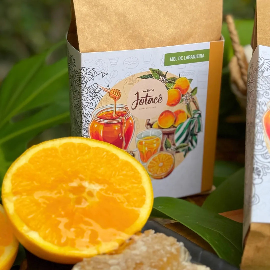 Close da embalagem do café Mel de Laranjeira da Fazenda Jotacê ao lado de laranja madura e mel, destacando um café especial naturalmente doce e aromático.
