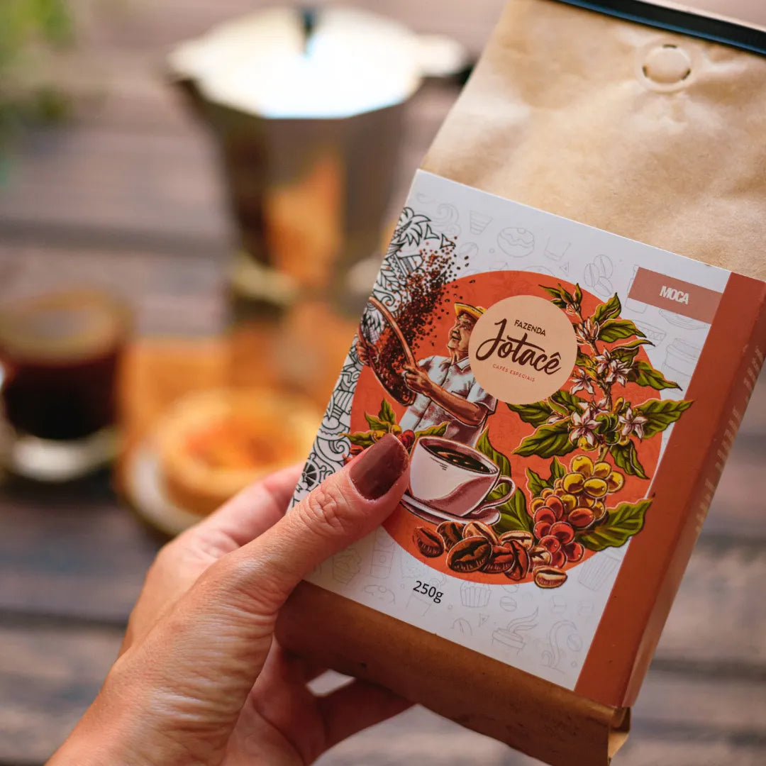 Mão segurando pacote de Café Microlote Moca com ilustração que remetem à cafeicultura
