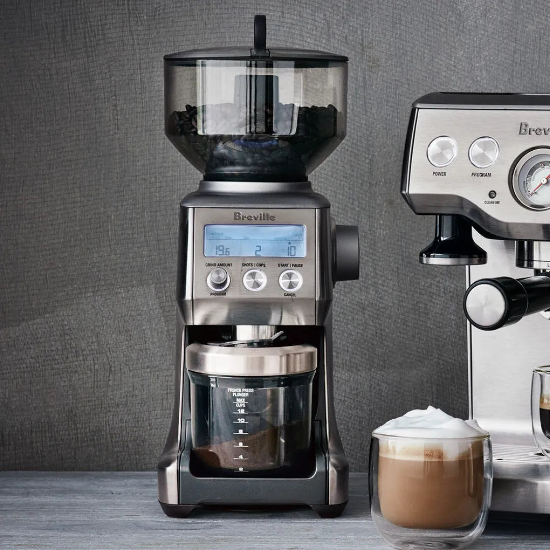 Moinho de café Breville ao lado de uma máquina de espresso e uma xícara de cappuccino, sobre um balcão.