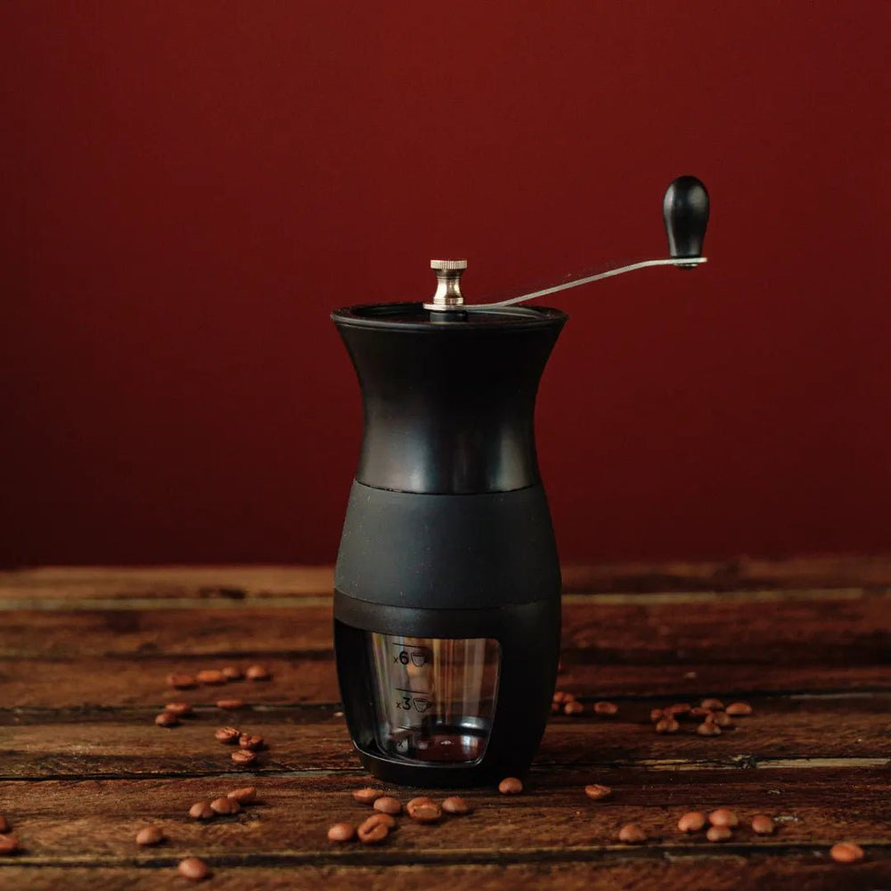 Moedor Manual Black de Café da Mimo Style com grãos de café ao redor sobre mesa de madeira.