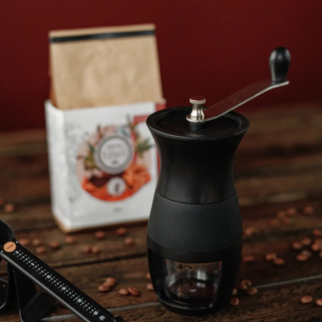 Moedor Manual Black para cafés da Mimo Style sobre mesa de madeira em primeiro plano e atrás uma embalagem desfocada do Kawá Caramelo 250g
