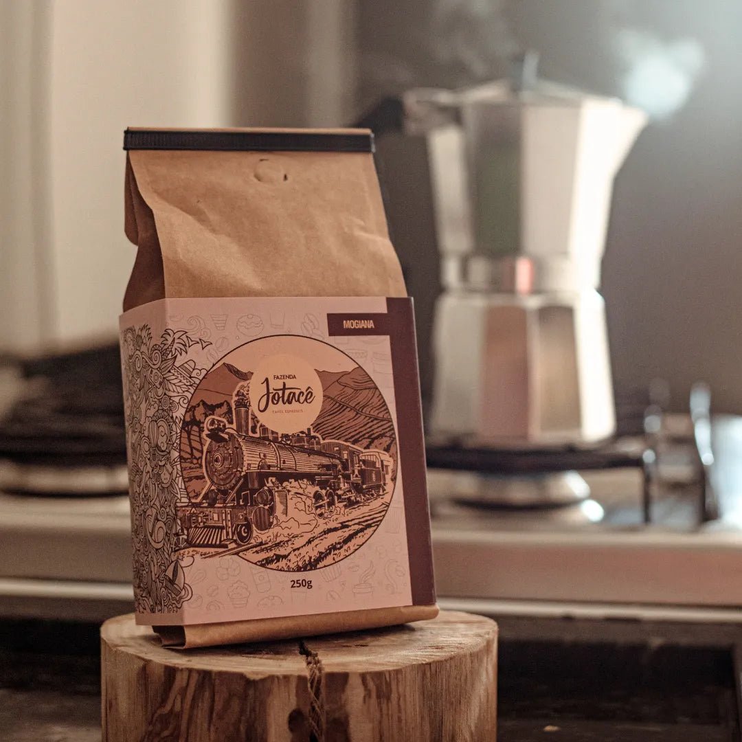 Pacote de Café Jotacê Mogiana em ambiente de cozinha com uma cafeteira Italiana ao fundo.