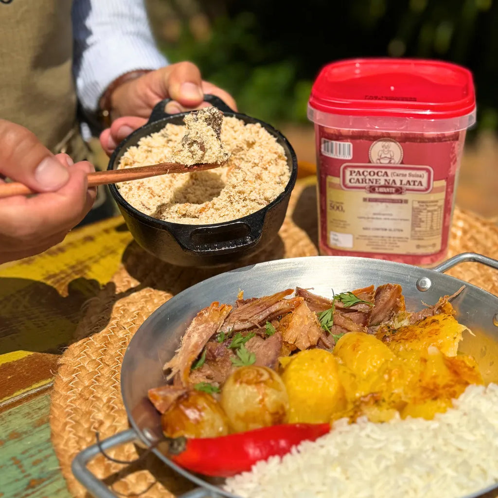 Paçoca de carne suína Xavante sendo servida com colher de pau, ao lado de prato com pernil suíno desfiado, batatas, arroz e pimenta, com o pote do produto ao fundo.
