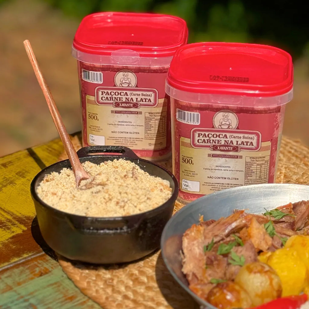 Dois potes de Paçoca Xavante 500g (carne suína) sobre mesa rústica, com panelinha cheia da paçoca e prato de carne de lata desfiada ao lado