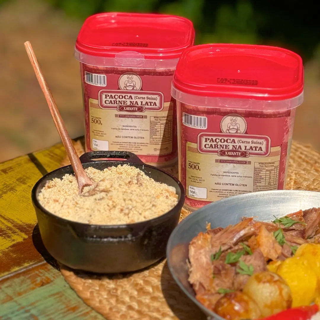 Dois potes de Paçoca Xavante 500g (carne suína) sobre mesa rústica, com panelinha cheia da paçoca e prato de carne de lata desfiada ao lado