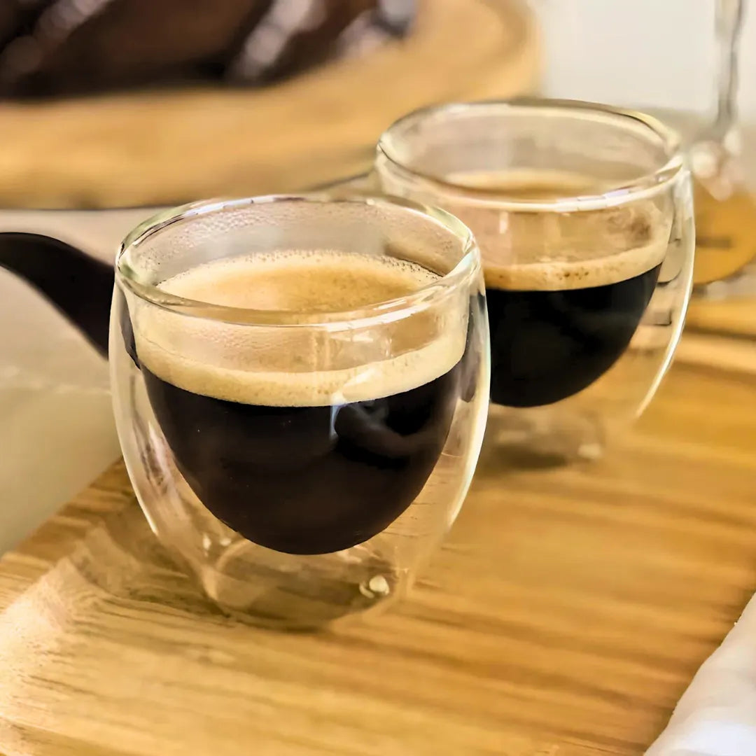 Duas xícaras de café expresso servidas em copos de vidro duplo sobre uma bandeja de madeira.