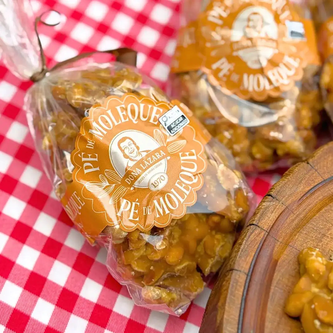 Close de pacote de Pé de Moleque Dona Lázara com rótulo laranja circular mostrando logotipo da marca desde 1970, amendoins caramelizados visíveis através da embalagem, sobre toalha xadrez vermelha