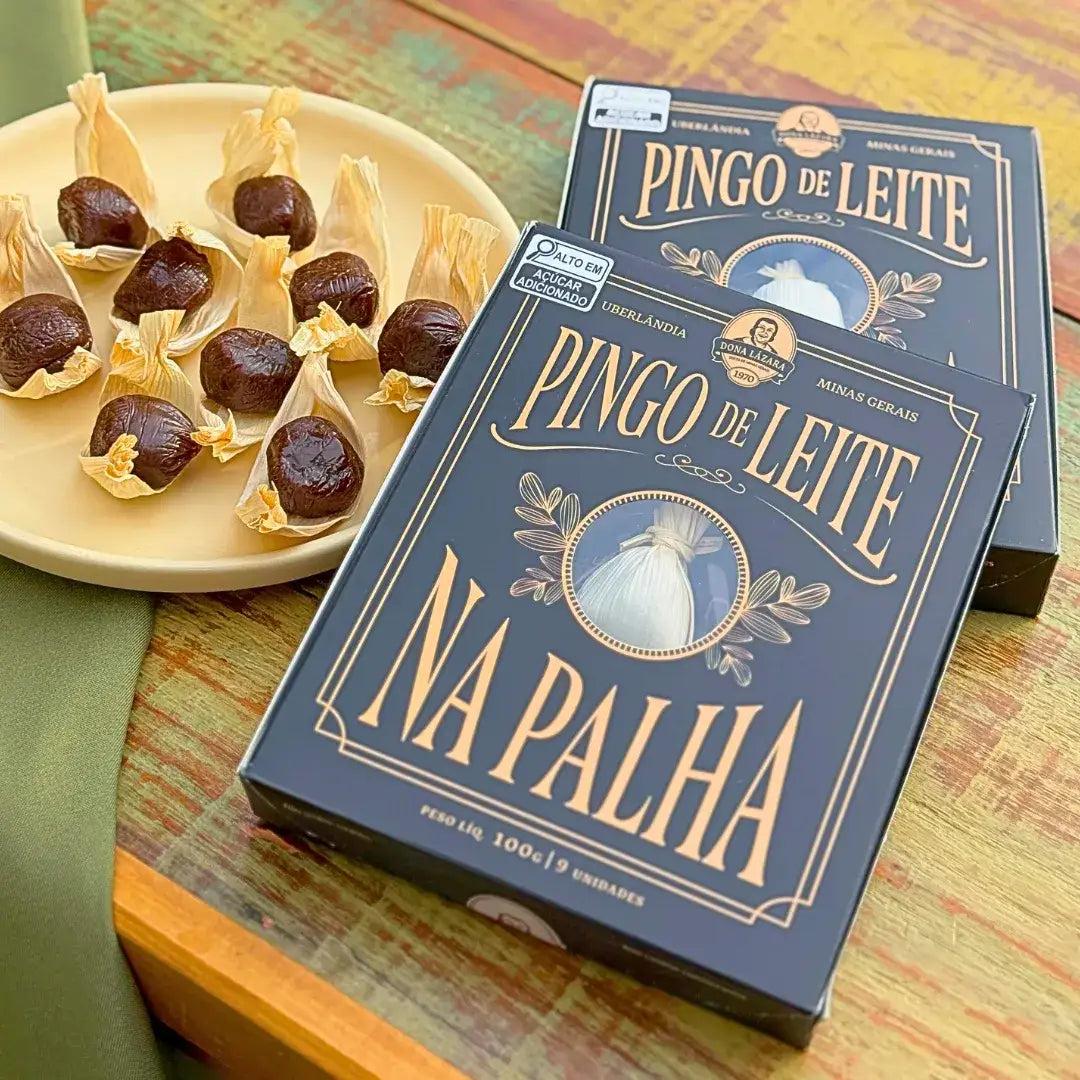 Caixa azul-escuro de Pingo de Leite na Palha Dona Lázara 100g sobre mesa rústica de madeira ao lado de prato com doces de leite abertos na palha de milho