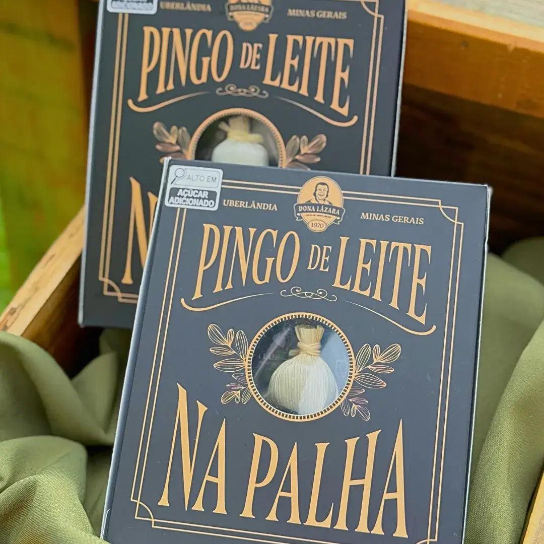 Duas caixas de Pingo de Leite na Palha Dona Lázara em azul-escuro com detalhes dourados sobre tecido verde, visor circular mostrando doce embrulhado em palha, marca de Uberlândia MG