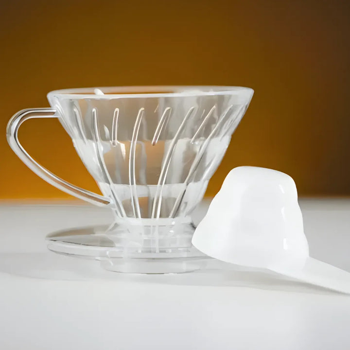 Porta filtro Cônico transparente da Extratto ao lado de colher dosadora sobre mesa branca e fundo desfocado