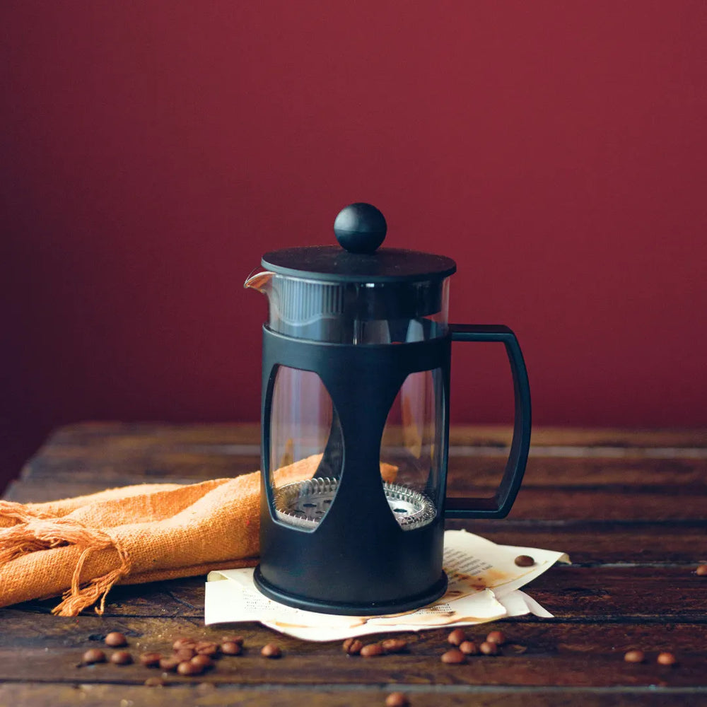 Prensa francesa Mimo Style preta com café fresco sobre pano e grãos de café ao redor.