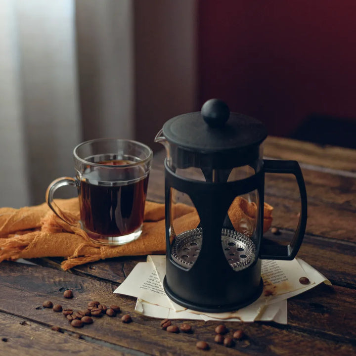 Prensa francesa preta ao lado de xícara de café e grãos de café sobre mesa de madeira.