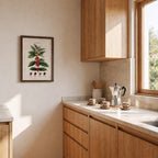 Quadro do pé de café emoldurado na parede de uma cozinha de marcenaria clara, ao lado da bancada com cafeteira italiana, xícaras e pote de utensílios.