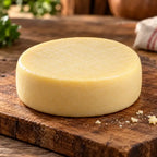 Peça inteira de queijo artesanal de leite cru, redonda e de casca amarela lisa, sobre tábua de madeira com farelos de queijo.