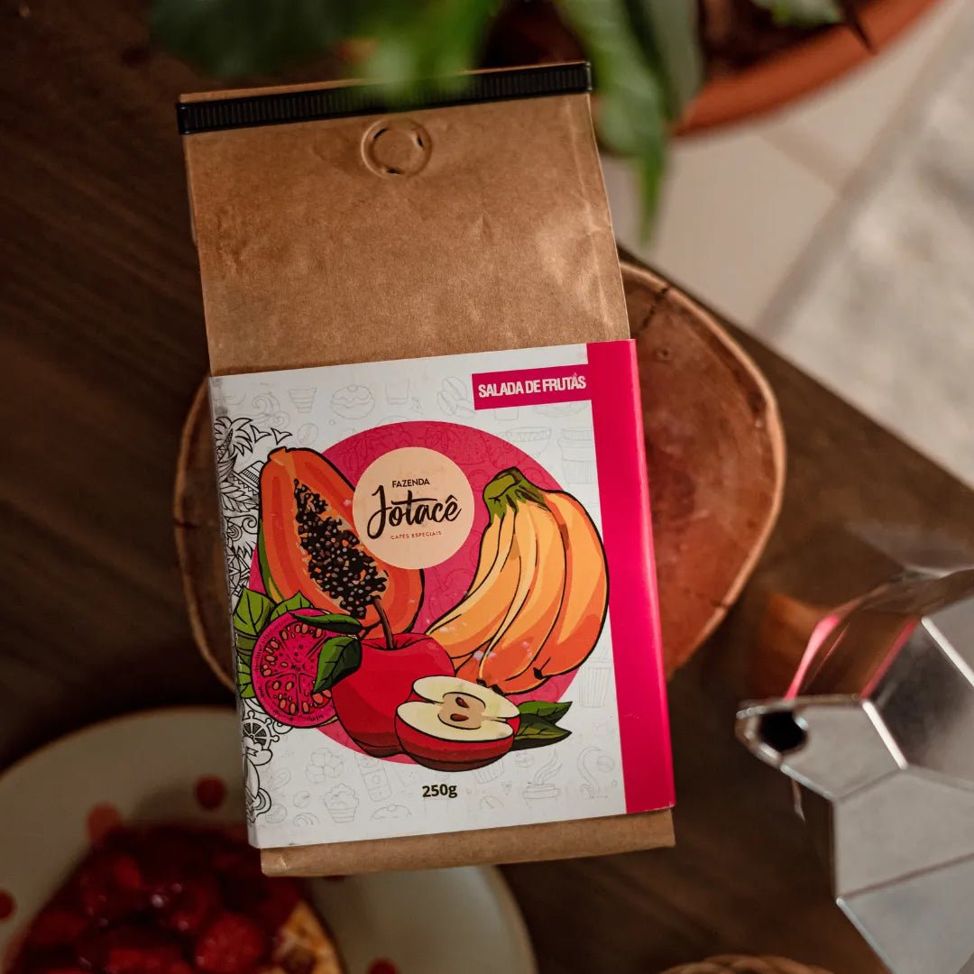 "Pacote de Café Jotacê Salada de Frutas sobre mesa de cozinha, ao lado de uma Cafeteira Italiana