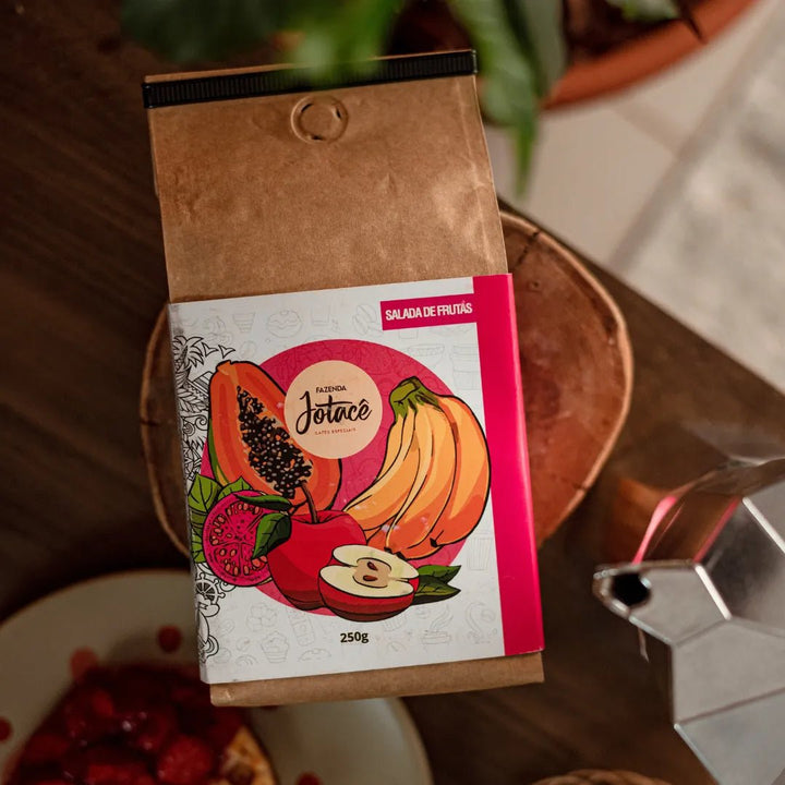 "Pacote de Café Jotacê Salada de Frutas sobre mesa de cozinha, ao lado de uma Cafeteira Italiana