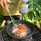 Torresmo Xavante sendo frito em panela de ferro sobre fogareiro, com a lata da carne na lata Xavante segura ao fundo no jardim.