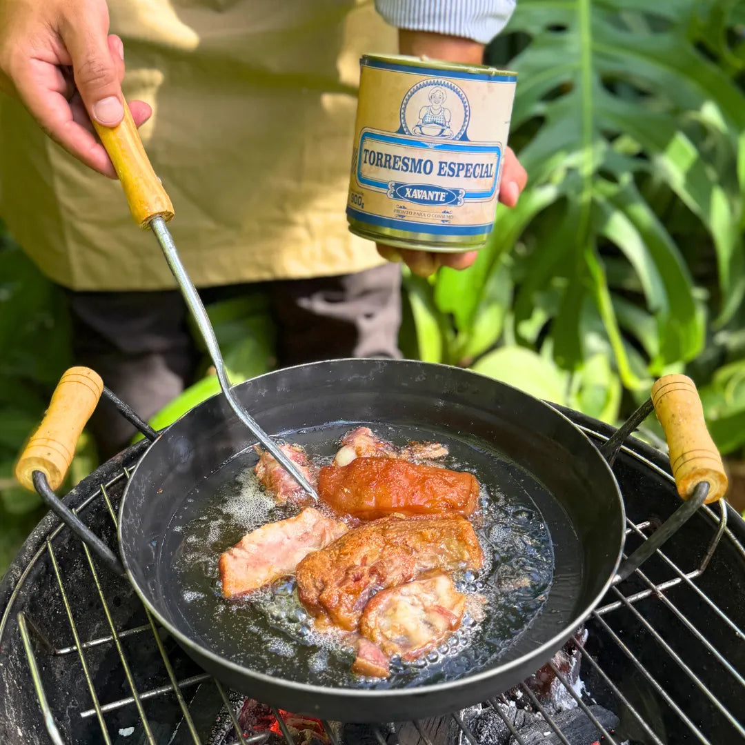 Torresmo Xavante sendo frito em panela de ferro sobre fogareiro, com a lata da carne na lata Xavante segura ao fundo no jardim.