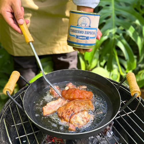 Torresmo Xavante sendo frito em panela de ferro sobre fogareiro, com a lata da carne na lata Xavante segura ao fundo no jardim.