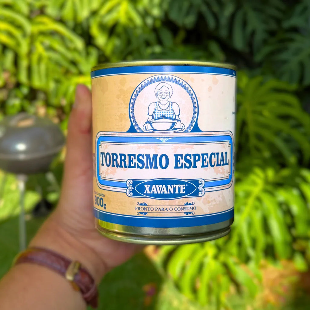 Lata fechada de Torresmo Especial Xavante 900g segurada na mão com fundo de folhagem tropical, embalagem azul e creme da carne de lata mineira.
