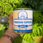 Lata fechada de Torresmo Especial Xavante 900g segurada na mão com fundo de folhagem tropical, embalagem azul e creme da carne de lata mineira.