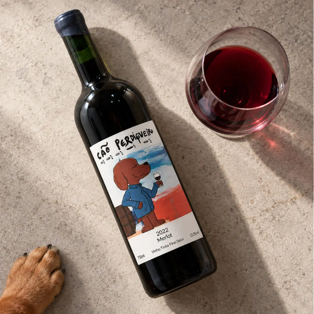 Vinho artesanal Cão Perdigueiro Merlot deitado ao lado de uma taça, visto de cima, com a pata de um cachorro