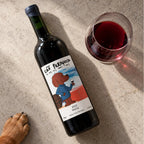 Vinho artesanal Cão Perdigueiro Merlot deitado ao lado de uma taça, visto de cima, com a pata de um cachorro