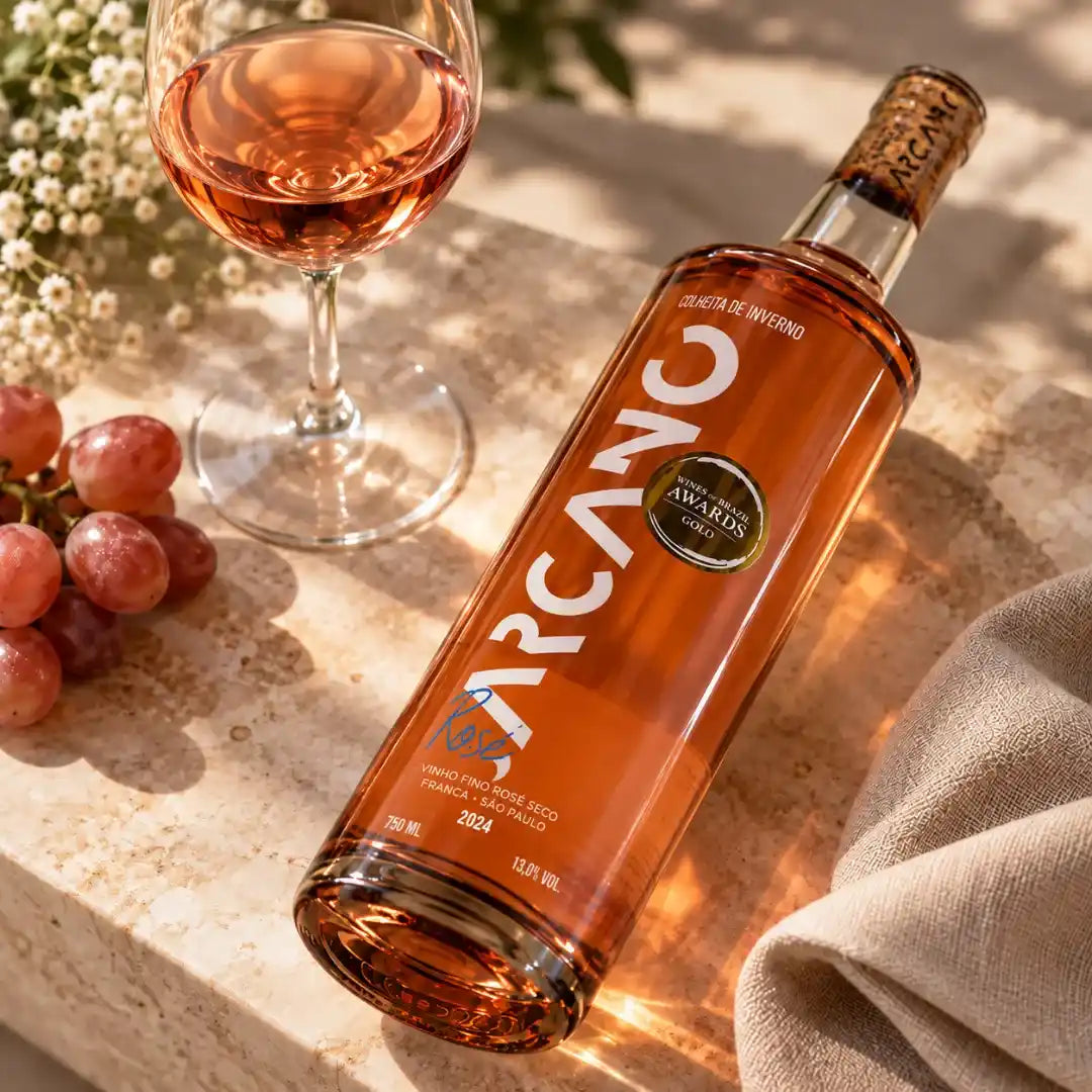 Garrafa de vinho Syrah rosé Arcano deitada sobre pedra clara, com taça servida, cacho de uvas rosadas e pano de linho sob luz de sol.