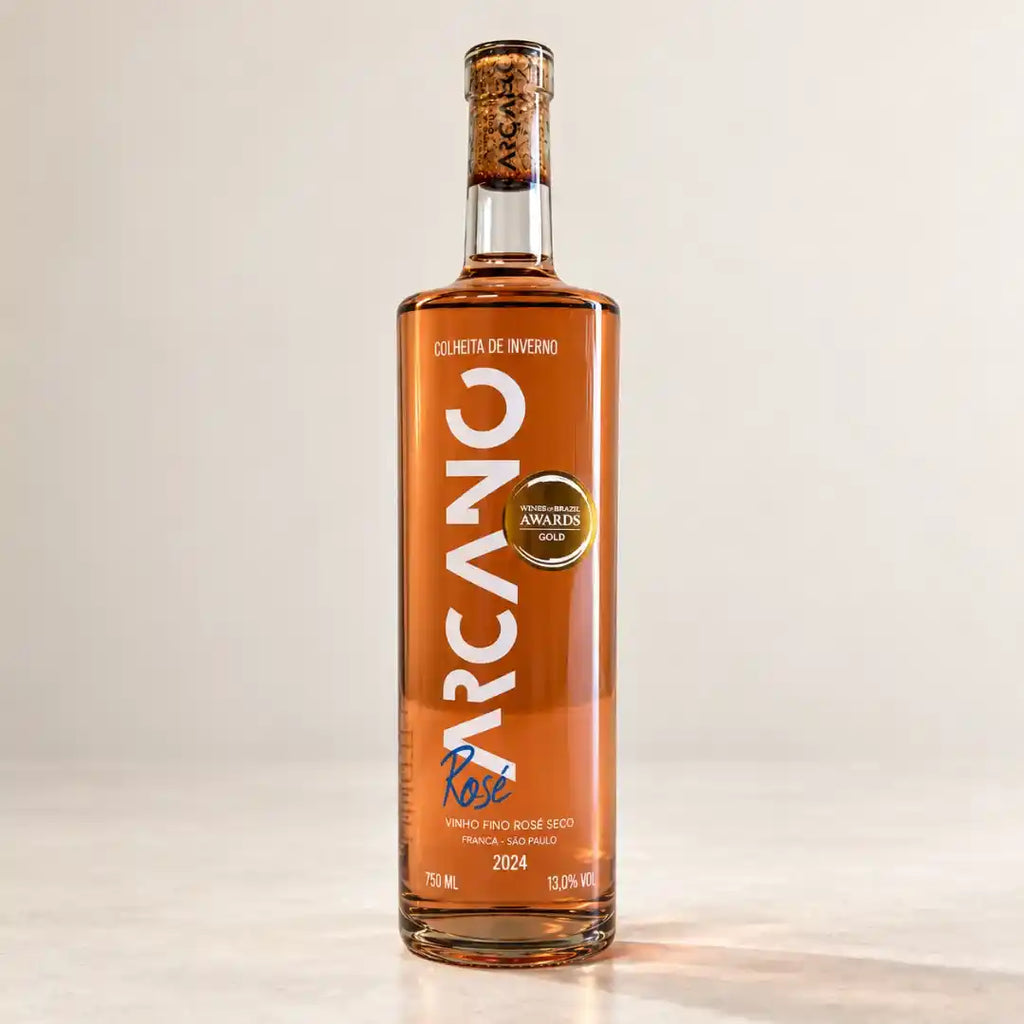 Garrafa do vinho rosé seco Arcano Syrah 750ml de frente, em fundo claro, com rótulo vertical branco, rolha à vista no gargalo e selo de ouro do Wines of Brazil Awards.