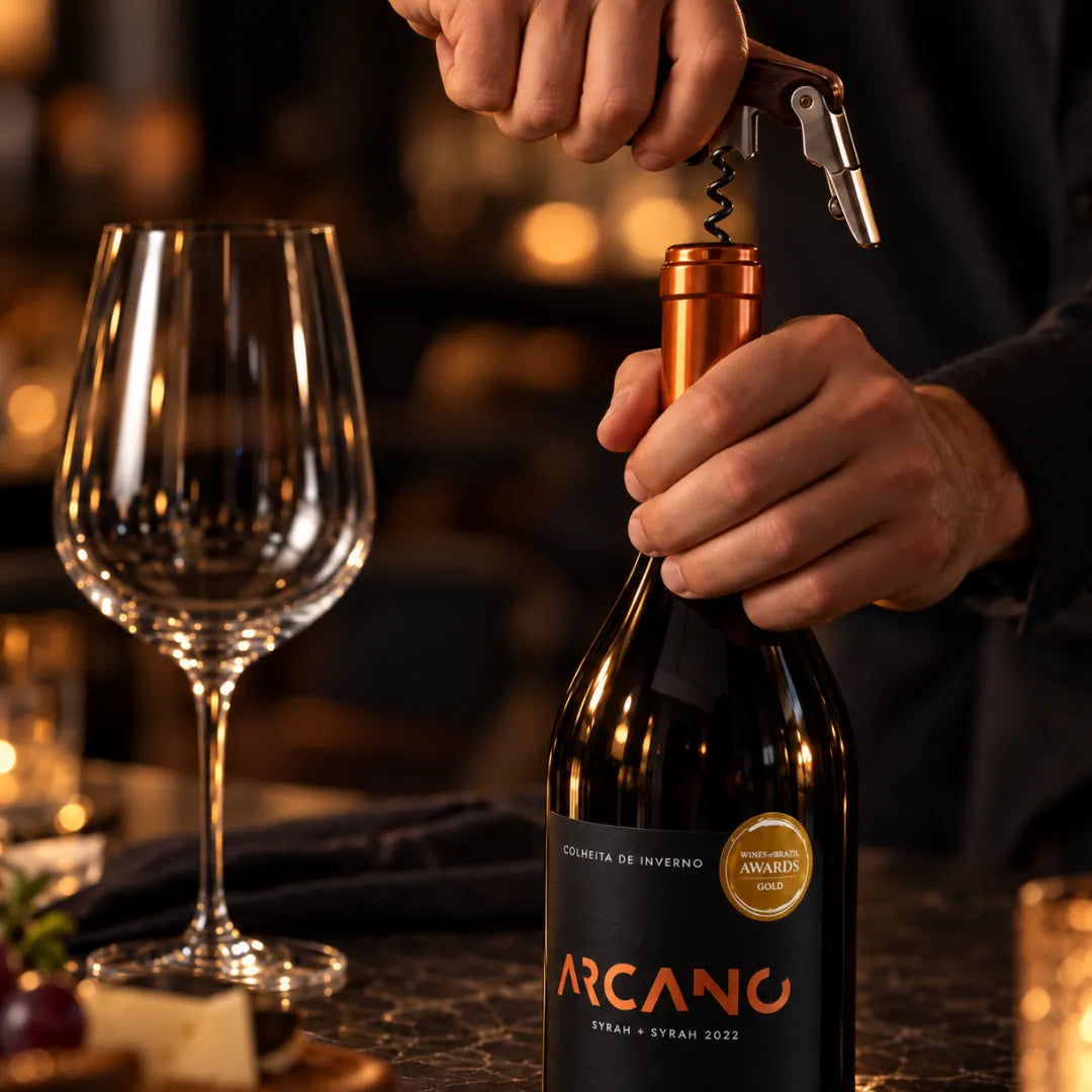 Mãos abrindo o vinho Syrah Arcano com saca-rolhas, com taça vazia, queijo e uvas sobre a mesa e luzes desfocadas ao fundo.
