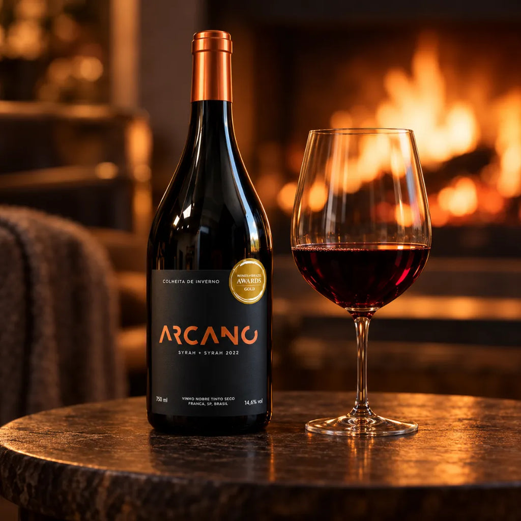 Vinho tinto Arcano Syrah ao lado de uma taça servida, sobre mesa de pedra em frente à lareira acesa.