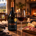 Arcano Syrah + Syrah, vinho nobre tinto seco de 14,6%, com taça de vinho tinto e tábua de queijo, salame, presunto cru e pão sobre mesa de madeira, com a serra ao fundo.