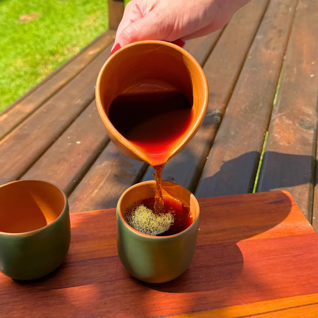 Mão despejando café de uma jarra de cerâmica dentro de uma xícara verde com interior laranja, sobre tábua de madeira ao ar livre com grama ao fundo.