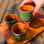 Mão com unhas vermelhas pegando uma xícara de cerâmica verde com interior laranja cheia de café, ao lado de outras duas na tábua de madeira ao sol.