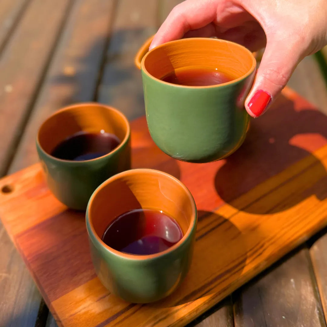 Mão com unhas vermelhas pegando uma xícara de cerâmica verde com interior laranja cheia de café, ao lado de outras duas na tábua de madeira ao sol.