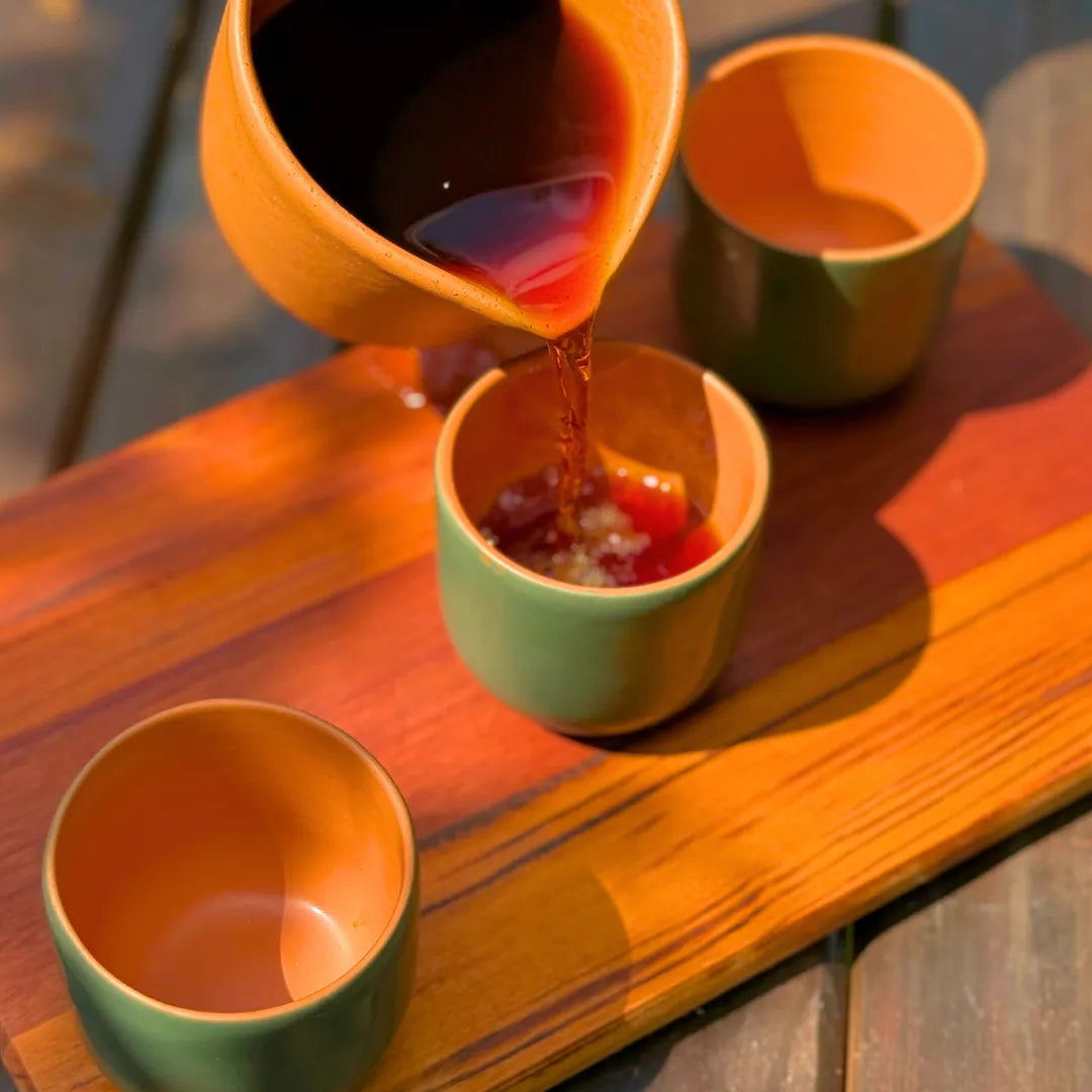 Jarra de cerâmica servindo café numa xícara verde com interior laranja sobre uma tábua de madeira ao ar livre, com outras duas xícaras da mesma cerâmica ao lado.