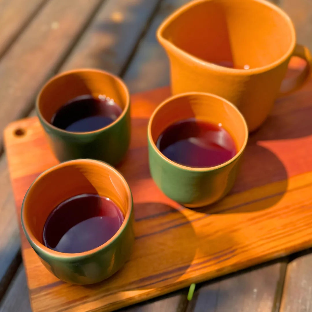 Três xícaras de cerâmica verde com interior laranja cheias de café e uma jarra da mesma cerâmica sobre uma tábua de madeira, vistas de cima na luz do sol.